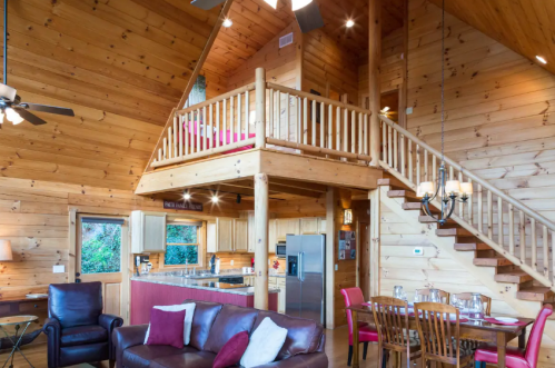 Cozy wooden cabin interior with a loft, featuring a living area, kitchen, and dining space. Warm, inviting atmosphere.