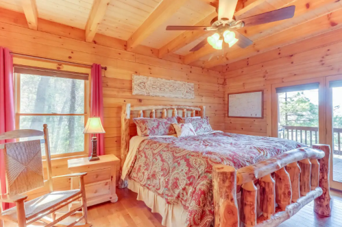 Cozy wooden bedroom with a large bed, rocking chair, and windows overlooking nature. Warm lighting and rustic decor.
