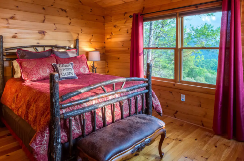 Cozy cabin bedroom with a wooden bed, red bedding, and a view of nature through a large window.