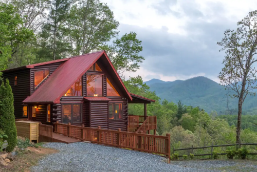 A cozy log cabin with a red roof, surrounded by trees and mountains, under a cloudy sky. Warm light glows from the windows.