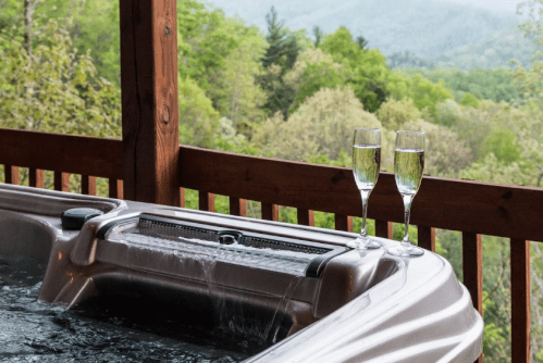 A hot tub on a deck with two glasses of champagne, overlooking a lush green landscape and mountains in the background.