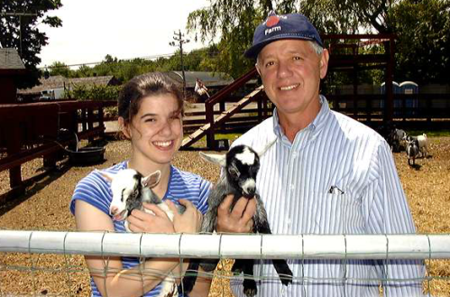 A man and a woman hold baby goats, smiling in a farm setting with a wooden fence and playground in the background.