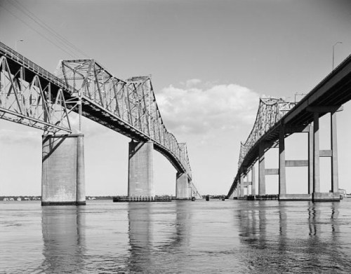 Black and white image of two large bridges spanning a calm body of water under a clear sky.