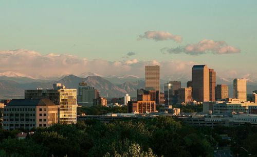 A city skyline with modern buildings against a backdrop of mountains and clouds during sunset.