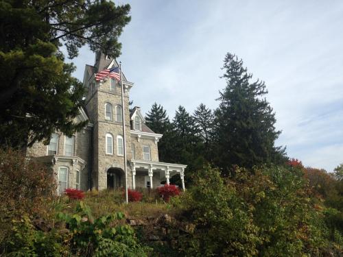 A tall, historic stone house with an American flag, surrounded by trees and colorful foliage under a cloudy sky.
