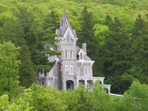 A tall, historic stone house with a pointed roof, surrounded by lush green trees and hills. An American flag is visible.