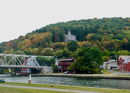 A scenic view of a hillside with a castle-like building, surrounded by colorful autumn foliage and a calm waterway below.