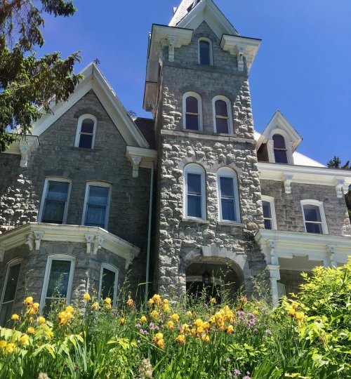 A large stone house with a tower, surrounded by vibrant yellow and purple flowers under a clear blue sky.