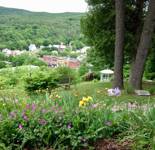 A scenic view of a town nestled in green hills, with colorful flowers in the foreground and a gazebo in the distance.