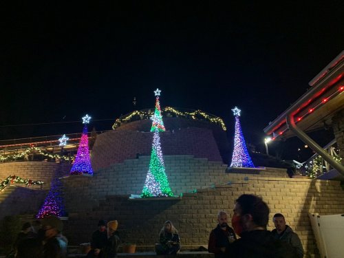 Colorful Christmas trees made of lights shine at night, surrounded by festive decorations and people enjoying the scene.