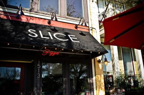 A storefront with a black awning reading "SLICE" and a red "Pizza" sign, surrounded by trees and outdoor seating.