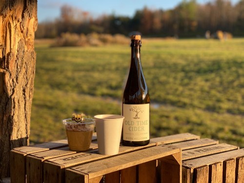 A bottle of Old Time Cider and a cup sit on a wooden crate, with a scenic field in the background during golden hour.