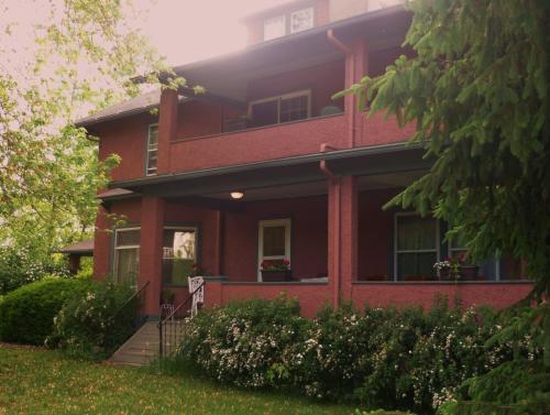 A two-story red house with a porch, surrounded by greenery and flowers, featuring large windows and a welcoming entrance.