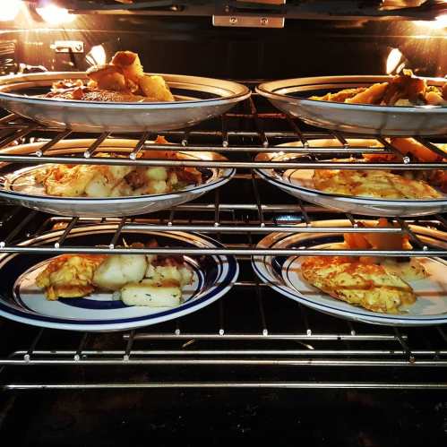 Plates of food in an oven, with various dishes on each shelf, illuminated by the oven's light.