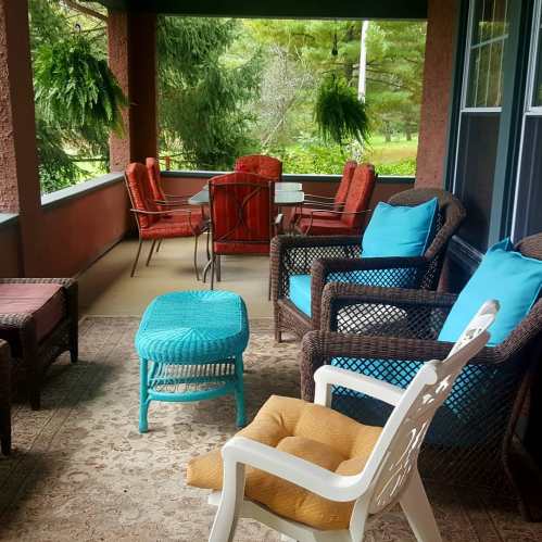 Cozy porch with wicker furniture, colorful cushions, and hanging plants, surrounded by greenery.