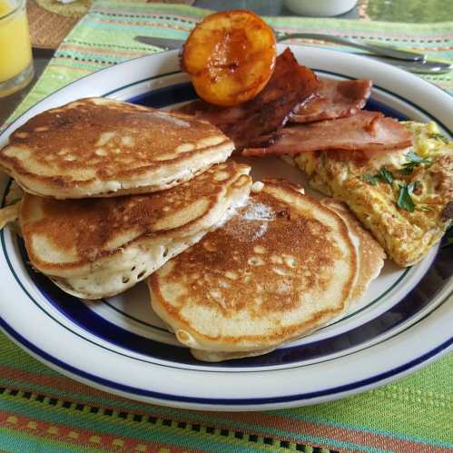 A plate of pancakes, bacon, and an omelet, with a grilled peach on the side, set on a colorful tablecloth.