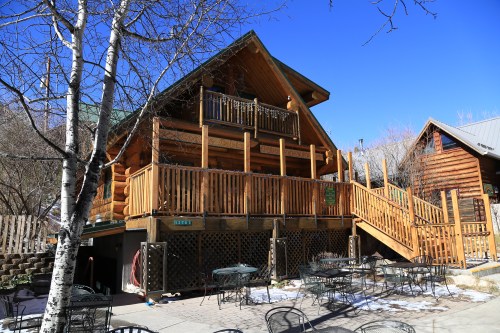 A rustic log cabin with a wooden deck, surrounded by trees and outdoor seating, under a clear blue sky.