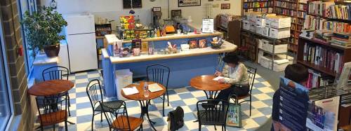 A cozy bookstore café with tables, a counter, and shelves filled with books. A person is writing at a table.