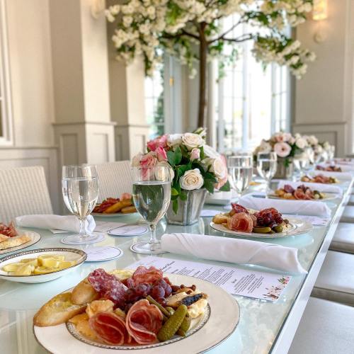 A beautifully set dining table with elegant plates of charcuterie, flowers, and glasses of water in a bright, airy room.