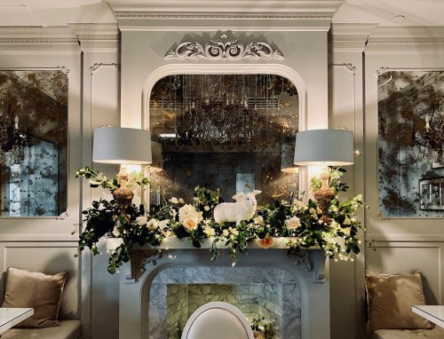 Elegant fireplace mantel adorned with flowers, greenery, and decorative lamps in a stylish interior setting.