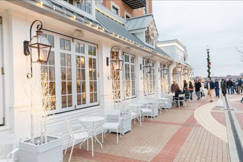 A charming outdoor café with white furniture and decorative lights, bustling with people along a brick walkway.