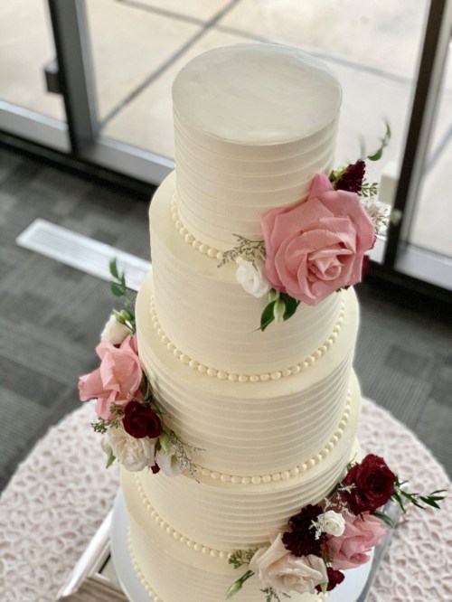 A tall, elegant wedding cake with smooth white frosting, adorned with pink and red roses and decorative pearls.