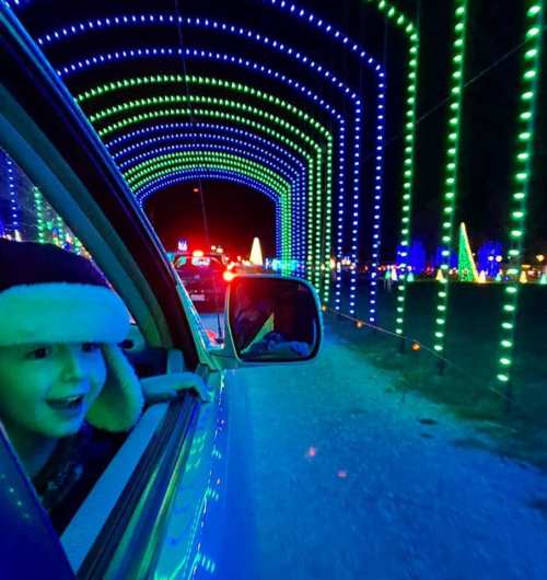 A child smiles from a car window, surrounded by colorful holiday lights in a festive display.