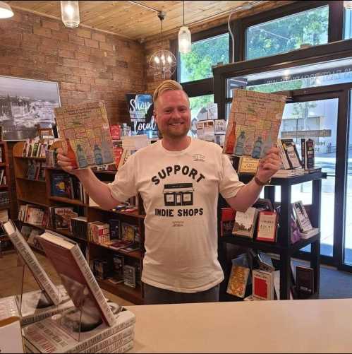 A smiling man in a "Support Indie Shops" t-shirt holds up two colorful books in a cozy bookstore.