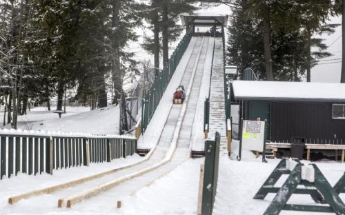 A snowy sledding hill with a steep ramp and a person sliding down on a sled. Trees and a building are in the background.