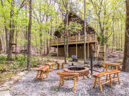 A cozy cabin in the woods surrounded by trees, with a fire pit and wooden benches in a gravel area.