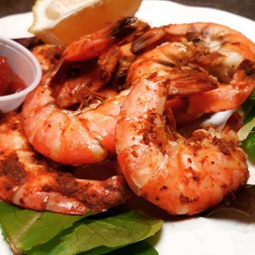 A plate of seasoned shrimp with a lemon wedge and a small cup of dipping sauce, garnished with fresh greens.