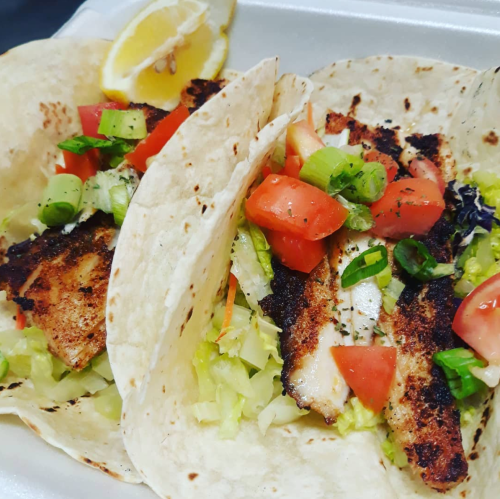 Two fish tacos filled with grilled fish, lettuce, tomatoes, and green onions, served with a lemon wedge.