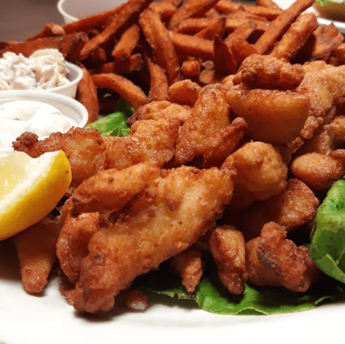 A plate of crispy fried chicken pieces, sweet potato fries, and dipping sauces, garnished with lemon and lettuce.