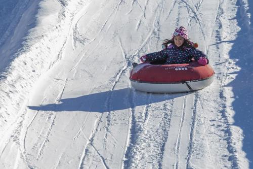 A child in a winter coat joyfully sleds down a snowy hill on a red inner tube.