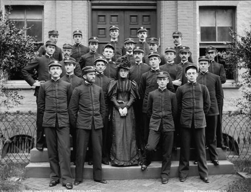 Group portrait of a naval academy class, featuring men in uniforms and a woman in a dress, posed in front of a building.