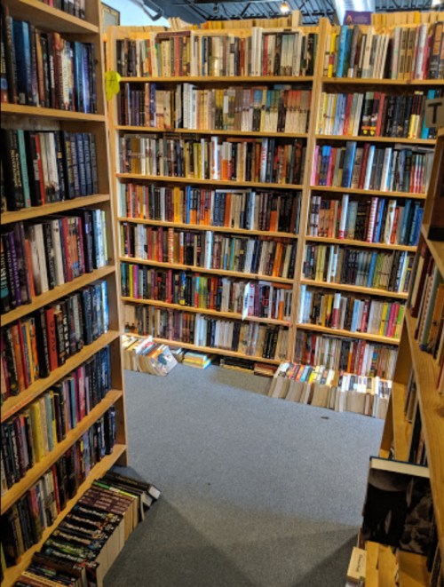 A cozy bookstore aisle lined with tall shelves filled with books, leading to a carpeted area with more books on the floor.