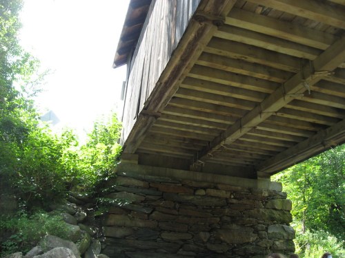 A wooden structure elevated on stone supports, surrounded by greenery and sunlight filtering through the trees.