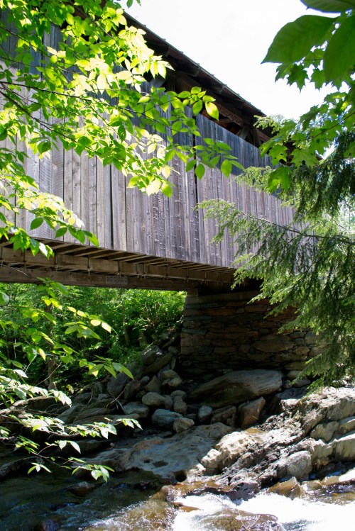 A wooden covered bridge spans a rocky stream, surrounded by lush green foliage and trees.