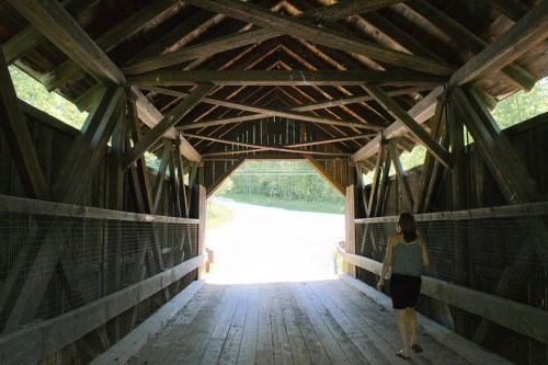 A person walks through a wooden covered bridge, with sunlight illuminating the exit and trees in the background.