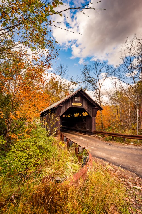 A rustic covered bridge surrounded by vibrant autumn foliage under a partly cloudy sky.