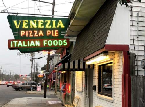 Neon sign for Venezia Pizza Pie, featuring "Italian Foods," outside a casual restaurant on a street corner.