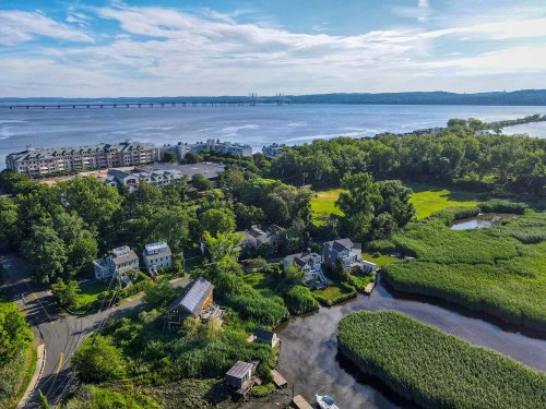 Aerial view of a riverfront area with lush greenery, houses, and a bridge in the distance under a clear blue sky.