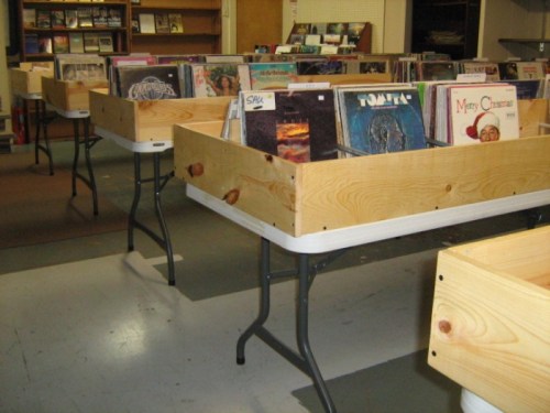 Wooden tables filled with vinyl records in a spacious room, with shelves of books in the background.