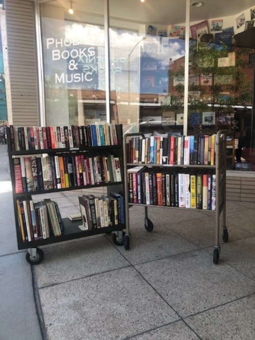Two book carts filled with various books, positioned outside a bookstore with large windows and a sunny atmosphere.