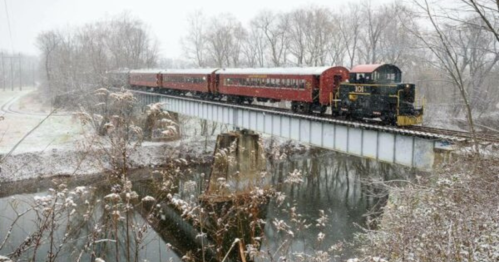 A red train crosses a snowy bridge over a river, surrounded by bare trees in a winter landscape.