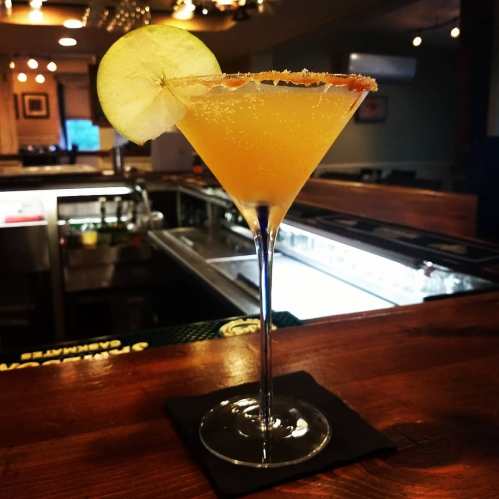 A vibrant cocktail in a tall glass, garnished with a slice of apple and a sugared rim, sits on a wooden bar.