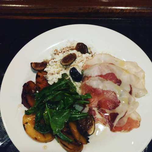 A plate with sliced cured meat, cottage cheese, sautéed peaches, and arugula, garnished with figs.