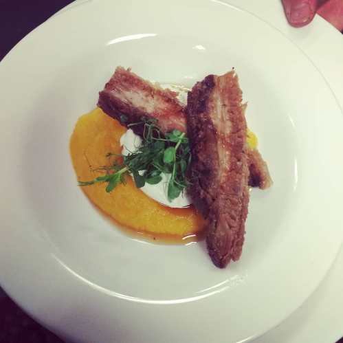 A plate featuring two pieces of crispy pork belly on a bed of creamy orange puree, garnished with microgreens.