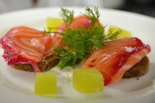 A plate featuring slices of salmon on dark bread, garnished with dill and green cubes, with a dollop of sauce.