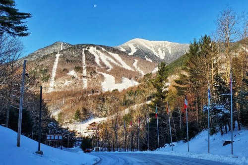 A snowy mountain landscape with ski trails, trees, and a clear blue sky, featuring a distant moon.